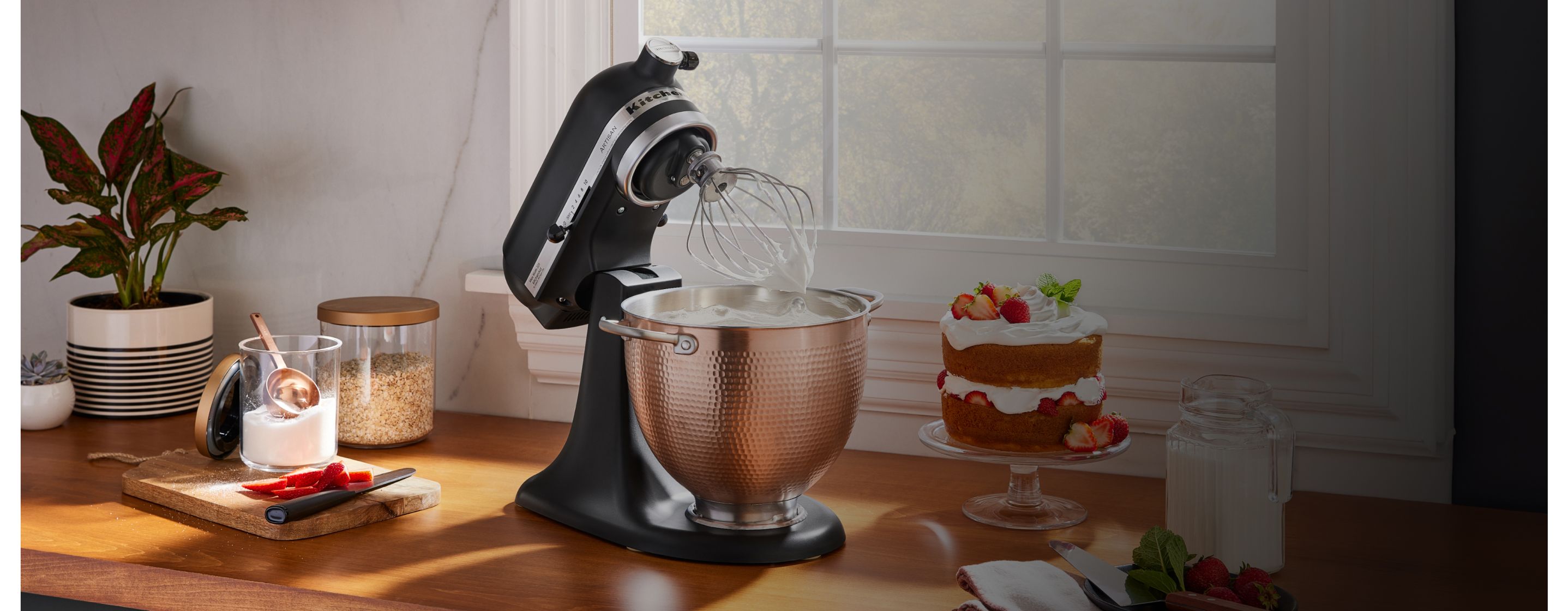 A black KitchenAid® stand mixer with a copper bowl on the counter, near a baked cake and other baking ingredients. 