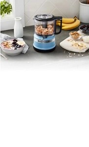 A blue chopper with nuts inside on a stone counter next to a bowl of oatmeal and a cutting board with fruit and other oatmeal toppings