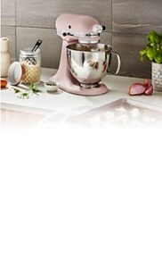 A pink KitchenAid® stand mixer with a stainless steel bowl on a white countertop next to flour, eggs, herbs and a baking tray