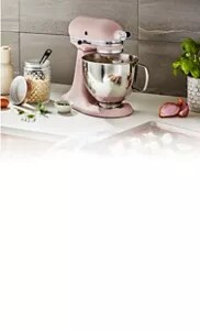 A pink KitchenAid® stand mixer with a stainless steel bowl on a white countertop next to flour, eggs, herbs and a baking tray