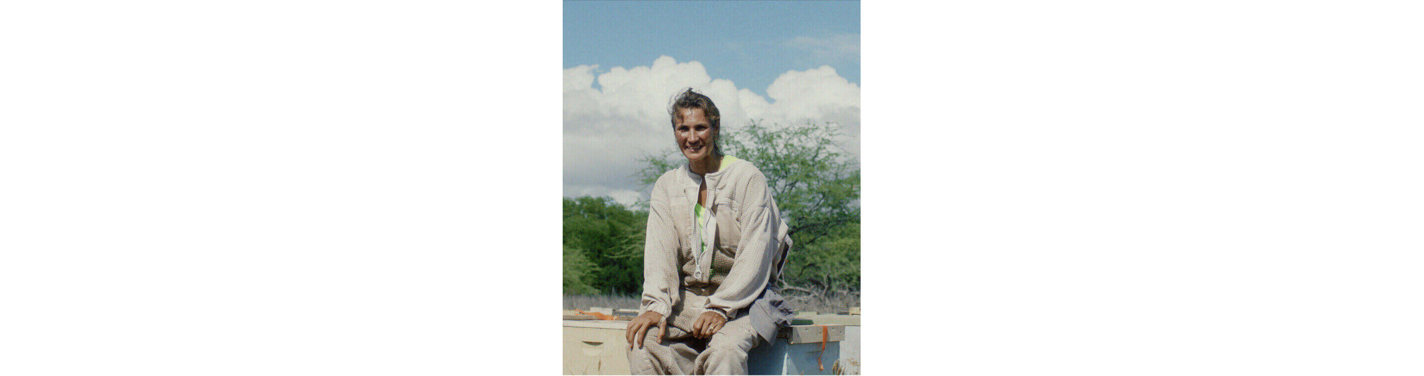 A woman in a beekeeping suit sitting down, smiling for the camera.